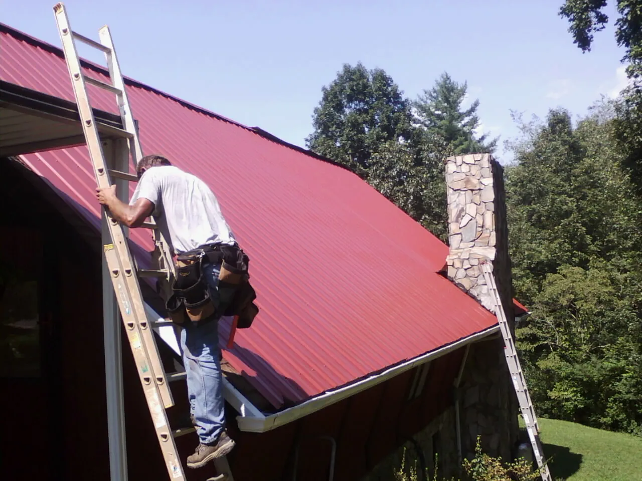 Roofer on a ladder beside a red metal roof and stone chimney — Soffit & Fascia Repair by Roofing Brothers in Whitehouse Station